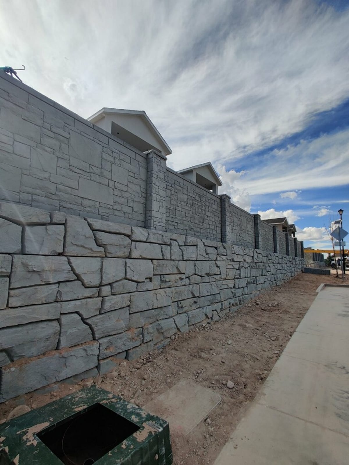 Decorative gray stacked-stone-style concrete block perimeter wall built by Roxor South on a Southern Utah jobsite, sidewalk in foreground under partial cloud sky